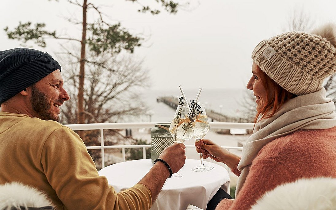 Paar stoßt im Winter auf Hotlelbalkon mit Ostsee-Aussicht mit Weißwein an